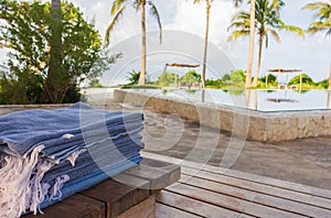 Stack of clean towels on wooden deck near swimming pool with palm trees. Lounge zone in tropical resort hotel. Empty hotel patio.