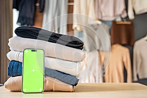 Stack of clothing prepared for resale with a smartphone displaying green screen for mockup use. Mobile apps simplify