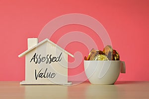 Stack of coins in a mug and toy house model with text ASSESSED VALUE