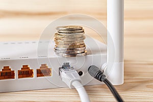 Stack of coins on the white Wi-Fi wireless router with connected cables on a wooden desk. Monthly payment of internet, pay bills