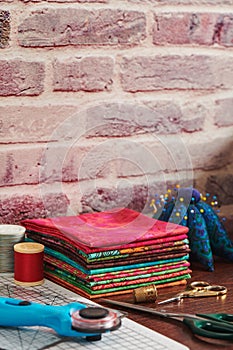 Stack of colorful fabrics on table surrounded by sewing accessories on brick wall background