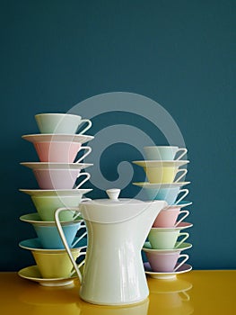 Stack of colorful vintage tea and coffee cups and tea pot on yellow table against petrol background. Vertical.