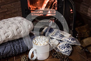 Stack of cozy knitted sweaters, handmade mittens and cup of coffee with marsh mallows on old wooden table, near burning