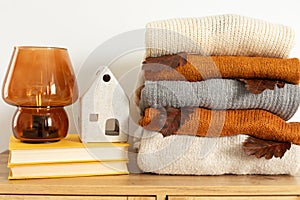 Stack of cozy knitted warm sweater with fall leaf, books, decorative broun lamp and ceramic house. Cozy hygge concept.