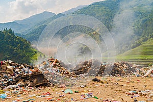 Stack of different types of large garbage dump, plastic bags, and trash burning near paddy rice terraces, agricultural fields of