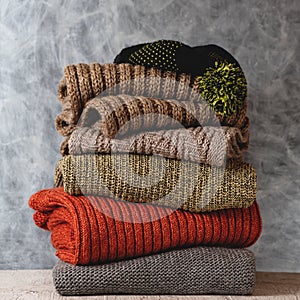 Stack of folded knitted wool women sweaters, scarf, hat with pompom on shelf against gray wall. Season of warm knitwear