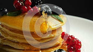 Stack of fresh fluffy pancakes decorated on top with forest berries rotating on plate and pouring honey syrup. Delicious