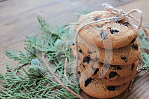 Stack of homemade chocolate chip cookies tied up with jute rope and evergreen twigs on wooden background.