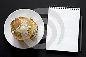 Stack of homemade pancakes with butter on white plate, blank notepad with pencil over black background, overhead view