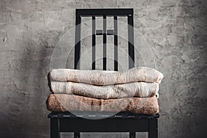 A stack of knitted warm cozy sweaters , on a chair by the gray wall. autumn