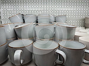 A stack of minimalist ceramic mugs displayed on a store shelf. Matte light-gray cups with subtle brown edging create a clean,