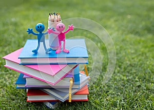 stack of multi-colored books lies on grass, on it are two funny souvenir pink and blue figures, set of pencils