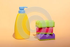 Stack of multi-colored dish wash sponges with bottle of soap on pastel orange background. Household cleaning scrub pad