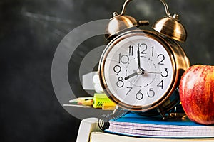 Stack of notebooks, workbooks, pads, pencils, supplies with alarm clock red apple on top. Black chalkboard. Back to school concept