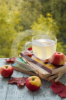 A stack of old books and drink with an apples and maple leaves on a wooden table near a wet window