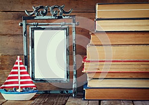 Stack of old books next to decorative sailing boat and blank frame wooden table. vintage filtered image