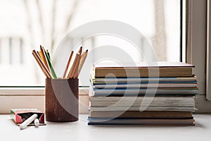 A stack of old books and several different colored pencils lie on the windowsill, a box of crayons lies on the side