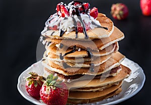 Stack of pancakes with strawberries, whip cream and chocolate syrup on a white plate on a black background.