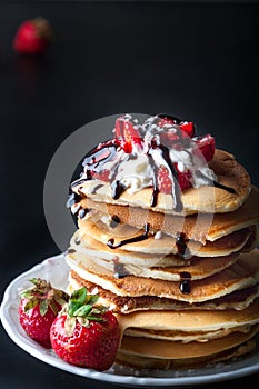 Stack of pancakes with strawberries, whip cream and chocolate syrup on a white plate on a black background.