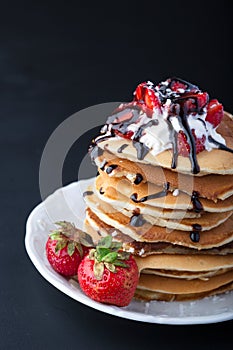 Stack of pancakes with strawberries, whip cream and chocolate syrup on a white plate on a black background.