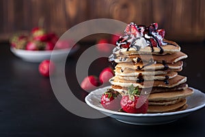 Stack of pancakes with strawberries, whip cream and chocolate syrup on a white plate on a black wooden background.