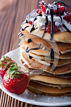 Stack of pancakes with strawberries, whip cream and chocolate syrup on a white plate on a wooden background.