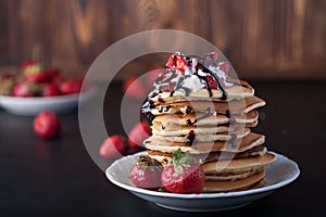 Stack of pancakes with strawberries, whip cream and chocolate syrup on a white plate on a wooden background.