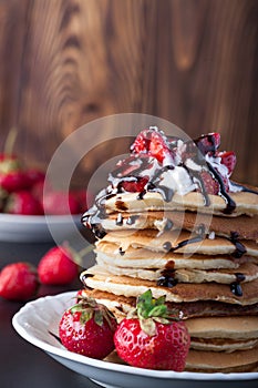 Stack of pancakes with strawberries, whip cream and chocolate syrup on a white plate on a wooden background.