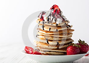 Stack of pancakes with strawberries, whip cream and chocolate syrup on a white wooden plate on a white woode background.