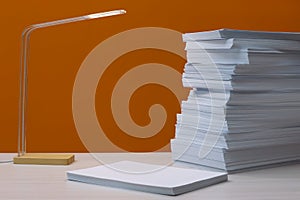 Stack of paper sheets and lamp on white wooden table near orange wall