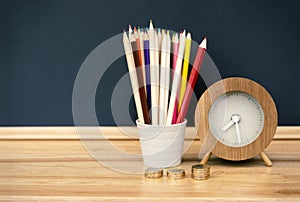 Stack of pound coins, colour pencils in white pot and wooden clock on wooden table with black broad background, Back to school con