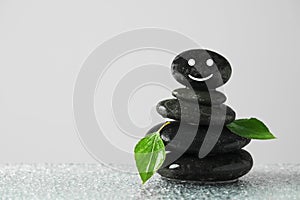 Stack of stones with drawn happy face, green leaves and water drops on table against grey background, space for text. Zen concept