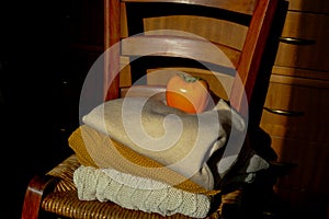 Stack of warm knitted sweaters in sunlight on wooden chairs across wooden dresser close-up and orange persimmon on the top. Warm c