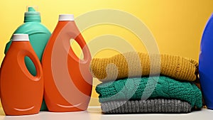 Stack of washed folded clothes and plastic orange large bottles with liquid detergent stand on a white table, yellow background. R