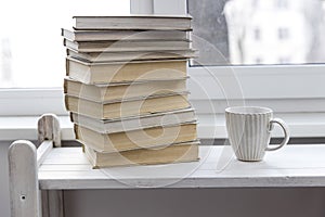 A stack of white-bound books is on table against the background of the window. The cup of tea is next to books. School