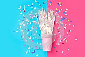 Stacked drinking paper cup striped straws colorful confetti scattered on bright duotone fuchsia blue background. Birthday
