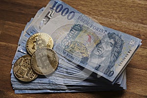 Stacks of 1000 Hungarian forints cash on brown wooden table. Next to it is a gold bitcoin digital cryptocurrency coin.