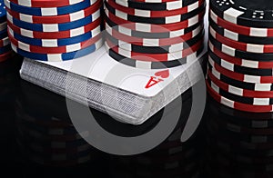Stacks of poker chips stand on a deck of playing cards and are reflected in a glossy black table close-up