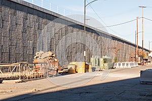 Staging area of road construction materials beside a concrete retaining wall and elevated highway