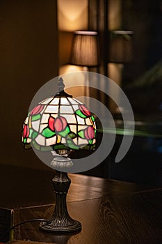 Stained glass lampshade with floral pattern. Glowing mosaic lamp closeup on dark blurry background. Vintage ornate table-lamp