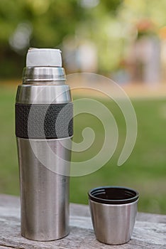 Stainless steel cup, mug with thermos stands on a wooden table in the forest on a green natural background. Thermos and