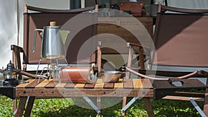 Stainless steel kettle, portable gas stove, bowl and vintage lanterns with outdoors wooden table set in front of tent on green