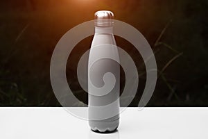 Stainless thermo bottle for water, tea and coffe, on white table. Dark grass background with sunlight effect. Thermos silver color