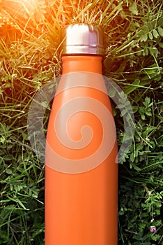 Stainless thermos water bottle, orange color. Mockup isolated on green grass background with sunlight effect. Glossy aluminum vacu