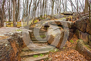 Stairs up among stone granite boulders in the park with trees without leaves. Autumn melancholic landscape on the background gray
