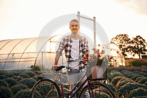 Standing, enjoying nature. Handsome man in casual clothes is with bicycle on the agricultural field near greenhouse