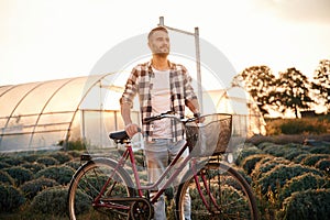 Standing, enjoying nature. Handsome man in casual clothes is with bicycle on the agricultural field near greenhouse