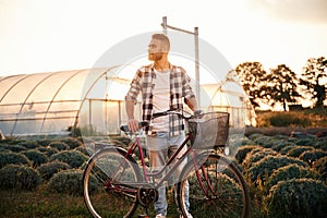 Standing, enjoying nature. Handsome man in casual clothes is with bicycle on the agricultural field near greenhouse