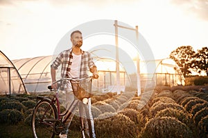 Standing, enjoying nature. Handsome man in casual clothes is with bicycle on the agricultural field near greenhouse