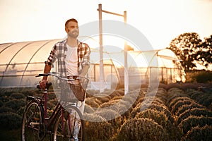 Standing, enjoying nature. Handsome man in casual clothes is with bicycle on the agricultural field near greenhouse